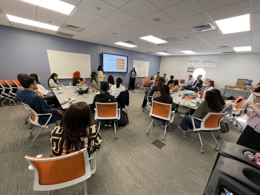 Two female students present their work in front of a room full of students and executives seated at three tables.